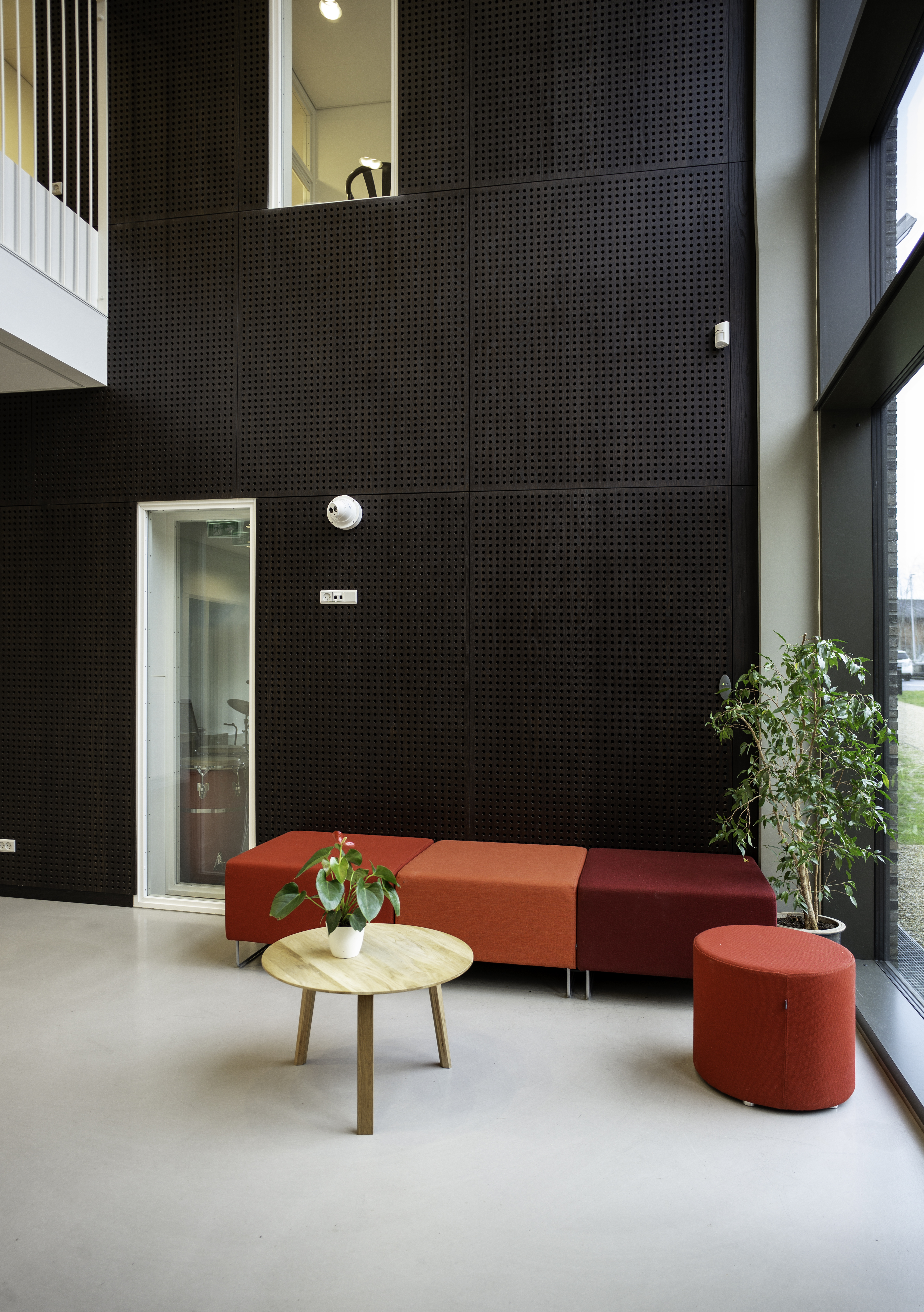 KulturRummet Thisted Musikskole - Mørke perforerede træpaneler på væg i atrium med røde siddemøbler